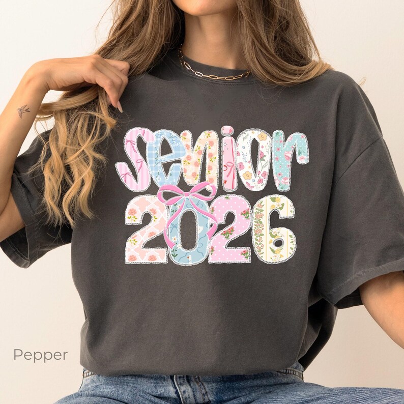 May include: Dark grey t-shirt featuring the text "Senior 2026" in a patchwork design. The letters and numbers are filled with floral and patterned fabrics in pink, blue, and white. A pink bow is between the words and numbers.