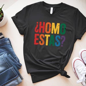 May include: Black t-shirt with the text "¿COMO ESTAS?" in rainbow colors. The shirt is made of a soft material and is likely a unisex design. The text is in a distressed font.