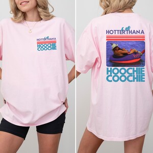May include: A light pink short-sleeve t-shirt featuring a graphic on the front left chest and a larger version on the back. The graphic includes red horizontal lines and the text HOTTER THAN A HOOCHIE COOCHIE with a photo of a person in a tube.