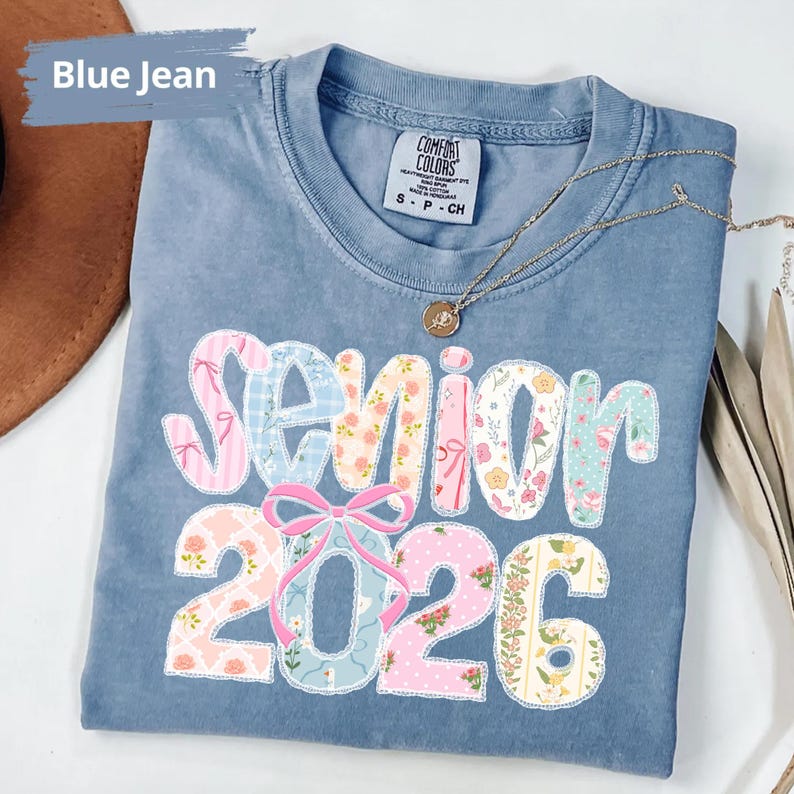 May include: A blue jean coloured t-shirt featuring the text "Senior 2026" in a patchwork design. The letters and numbers are decorated with floral and patterned fabrics. A gold necklace with a pendant is draped across the shirt.