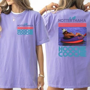 May include: A light purple short-sleeve t-shirt featuring a graphic on the front left chest and a larger version on the back. The graphic displays the text HOTTER THAN A HOOCHIE COOCHIE with red and teal lines and a photo of a person in a tube.
