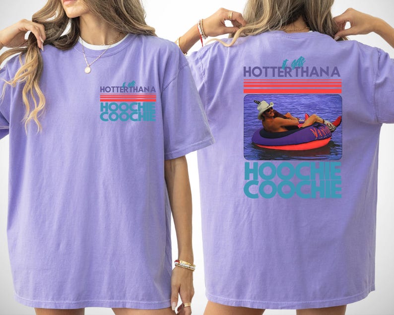 May include: A light purple short-sleeve t-shirt featuring a graphic on the front left chest and a larger version on the back. The graphic displays the text HOTTER THAN A HOOCHIE COOCHIE with red and teal lines and a photo of a person in a tube.