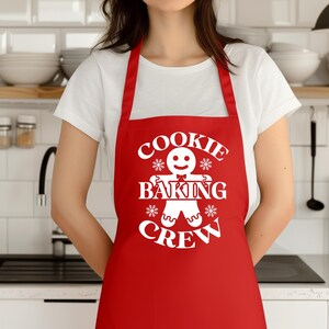May include: A red apron with the words "COOKIE BAKING CREW" in white, along with a gingerbread man graphic. The apron is being worn by a person in a white t-shirt.