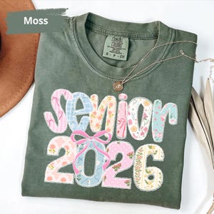 May include: A moss green t-shirt featuring the text "Senior 2026" in a patchwork design. The letters and numbers are decorated with floral and pastel patterns. A gold necklace with a pendant is draped across the shirt.