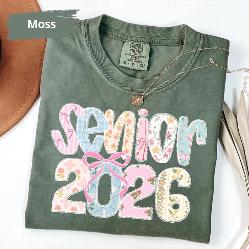 May include: A moss green t-shirt featuring the text "Senior 2026" in a patchwork design. The letters and numbers are decorated with floral and pastel patterns. A gold necklace with a pendant is draped across the shirt.