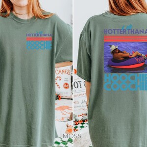 May include: A sage green short-sleeve t-shirt featuring a graphic on the front left chest and a larger version on the back. The graphic includes red horizontal lines and the text "HOTTER THAN A HOOCHIE COOCHIE" with a photo of a person in a tube.