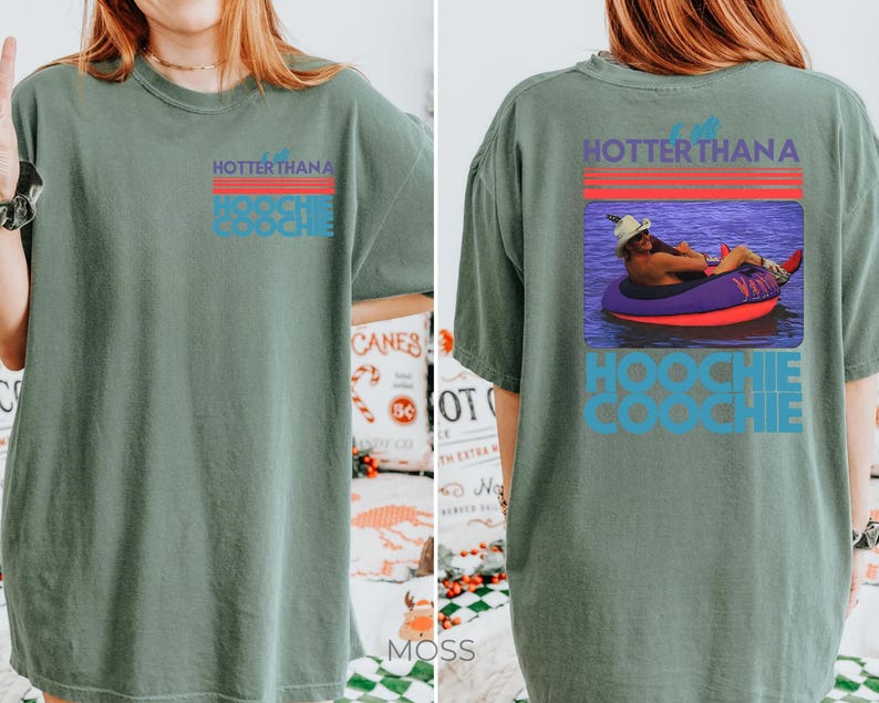 May include: A sage green short-sleeve t-shirt featuring a graphic on the front left chest and a larger version on the back. The graphic includes red horizontal lines and the text "HOTTER THAN A HOOCHIE COOCHIE" with a photo of a person in a tube.