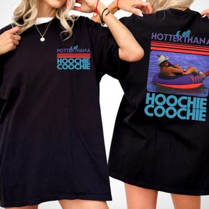 May include: A black t-shirt featuring a graphic print on the front and back. The front shows the text "IT GETS HOTTERTHANA HOOCHIE COOCHIE" in blue and red. The back displays the same text with a photo of a person in a tube on water.