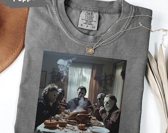 May include: Pepper-colored t-shirt with a graphic of a dinner scene featuring characters in masks. The shirt has a crew neck and short sleeves. A silver necklace with a pendant is draped on the shirt.