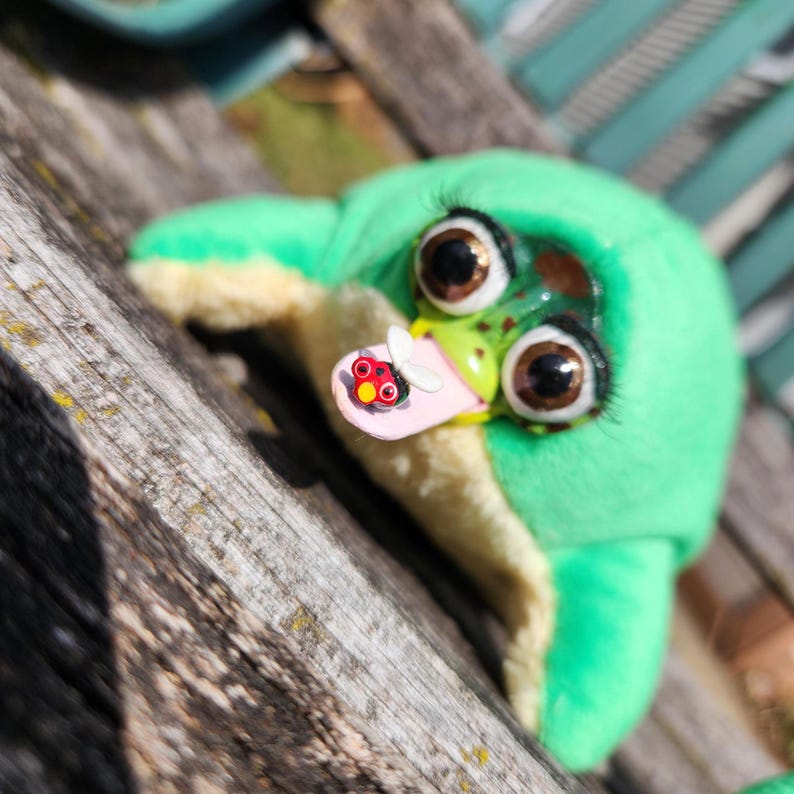 May include: A handmade plush frog with large, expressive eyes and a light green body. The frog has a pink tongue with a ladybug and a white butterfly attached. The frog is resting on a wooden surface.