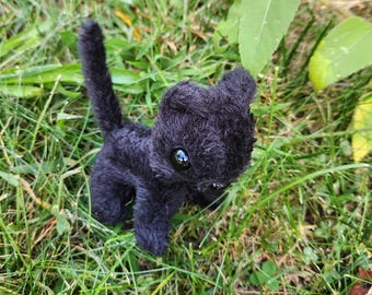May include: A small, black plush cat toy with large, reflective eyes and a fluffy tail. The cat is posed in a playful stance, set against a backdrop of green grass and foliage. The toy is made of a soft, textured material.