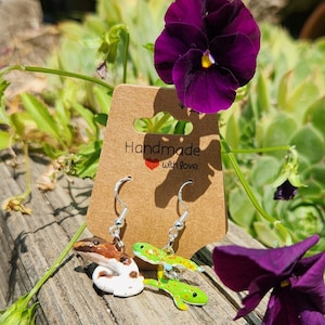 May include: A pair of dangle earrings featuring a green gecko and a brown and white gecko. The earrings are on a brown card with the text "Handmade with love".