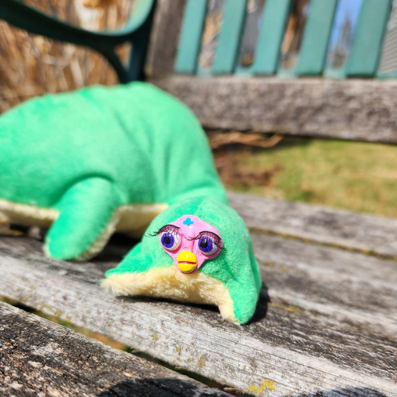 May include: A plush toy in the shape of a green creature with a long neck, lying on a wooden bench. The creature has large eyes with purple eyelashes, a pink face, and a yellow beak. The toy is a unique, handmade item.