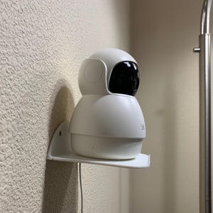 Puede incluir: Una cámara de seguridad blanca con una lente negra está montada en un estante blanco sujeto a una pared texturizada. La cámara tiene un diseño redondeado y está colocada cerca de una lámpara plateada.