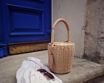 May include: A woven wicker handbag with a rounded shape and a handle, next to a white shirt and sunglasses. The bag has a tassel and is sitting on a concrete step in front of a blue door.