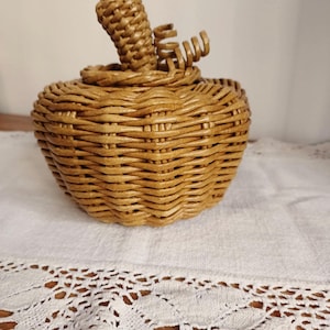 May include: A light brown woven wicker container shaped like a pumpkin. The object features a rounded body with a textured, interlaced reed pattern, a circular lid, and a central stem with a small, curled decorative vine detail on the top.