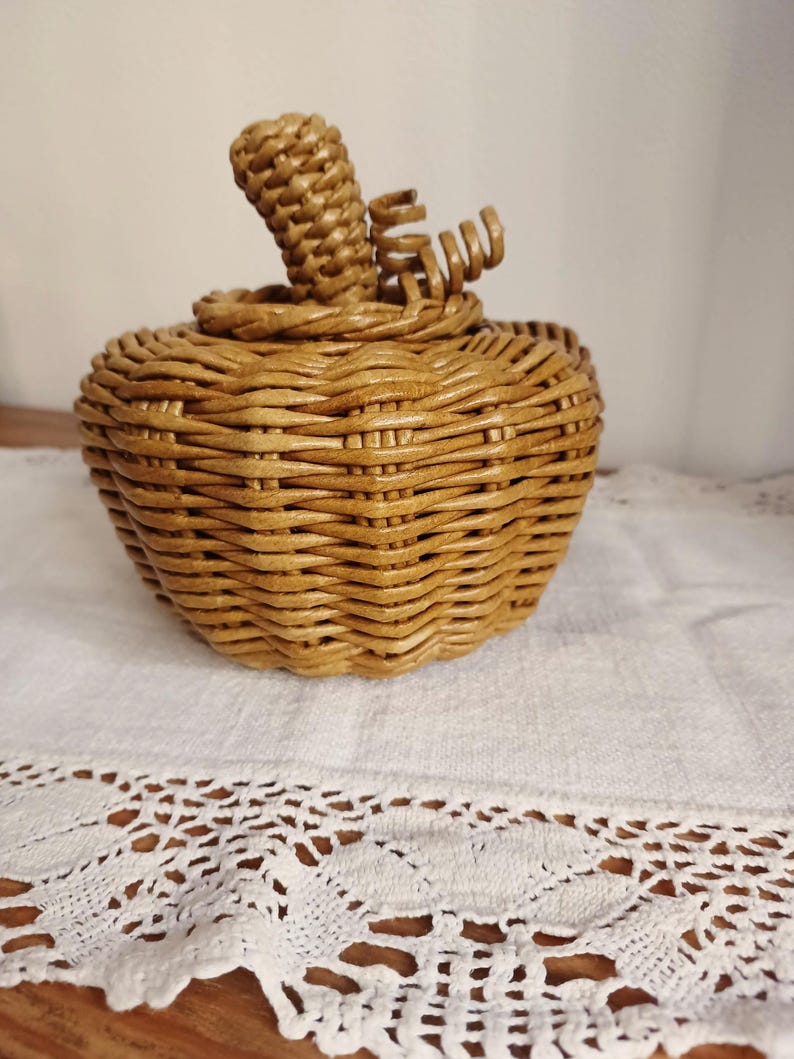 May include: A light brown woven wicker container shaped like a pumpkin. The object features a rounded body with a textured, interlaced reed pattern, a circular lid, and a central stem with a small, curled decorative vine detail on the top.
