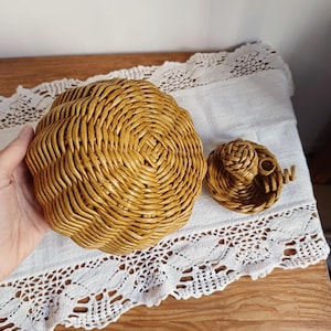 May include: A round, golden-brown woven basket with a textured, spiraled pattern and a matching separate lid featuring a small, intricate braided handle. The basket is constructed from coiled material with a glossy, uniform finish.