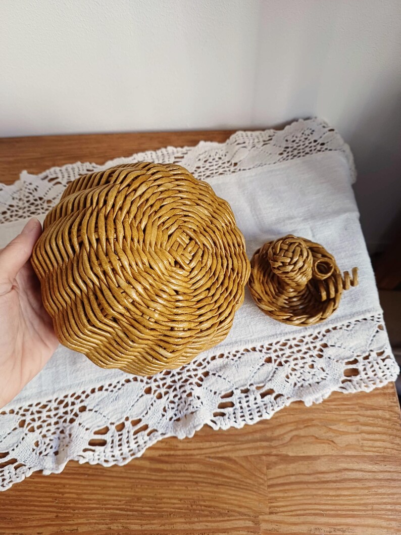 May include: A round, golden-brown woven basket with a textured, spiraled pattern and a matching separate lid featuring a small, intricate braided handle. The basket is constructed from coiled material with a glossy, uniform finish.