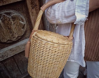 May include: A woven wicker handbag with a rounded shape and a light brown leather handle. The bag features a matching tassel and is held by a person wearing a white shirt and brown sweater. The bag is approximately 10 inches tall.