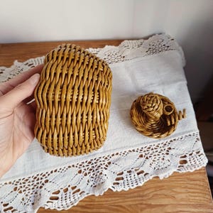 May include: A golden-brown woven wicker basket with a matching lid. The basket features a vertical weave pattern and a rounded shape. The lid is separate and includes a small, decorative woven knob on top, showcasing a detailed, hand-crafted texture.