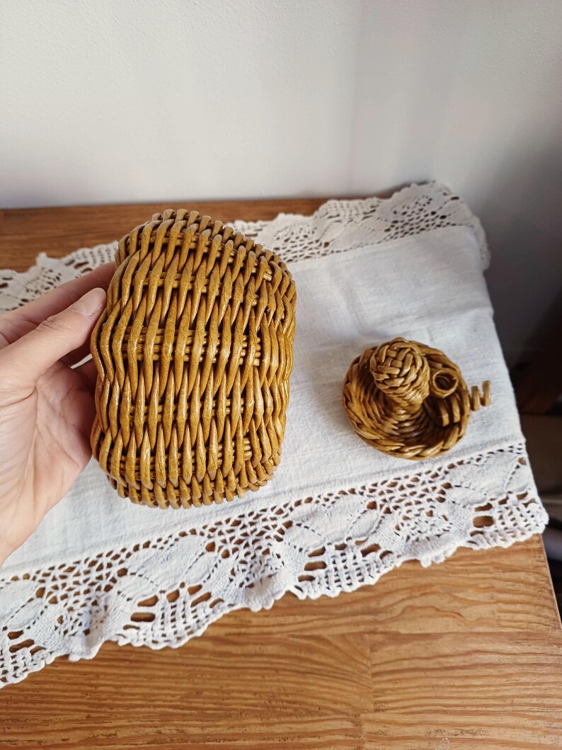 May include: A golden-brown woven wicker basket with a matching lid. The basket features a vertical weave pattern and a rounded shape. The lid is separate and includes a small, decorative woven knob on top, showcasing a detailed, hand-crafted texture.