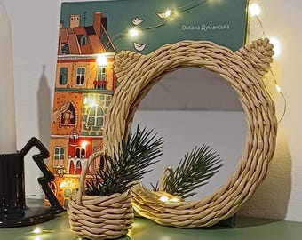 May include: A woven wicker mirror with a round shape and a light brown color. The mirror is decorated with a string of fairy lights and a small woven basket with a green plant. The mirror is sitting on a shelf with a book titled "Oksana Dumanska" behind it.