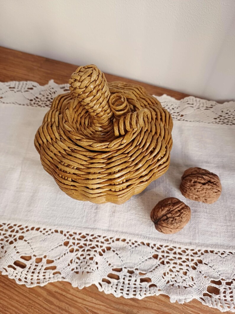 May include: A woven pumpkin-shaped basket with a golden-brown finish, featuring a coiled stem and two curled tendrils on top. Two whole walnuts are positioned next to the basket, which is constructed from tightly coiled, smooth material.
