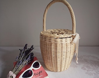 May include: A woven wicker basket with a rounded body and a curved handle. The basket is light brown and has a lid. Beside the basket are white sunglasses, a book, and a bundle of lavender.