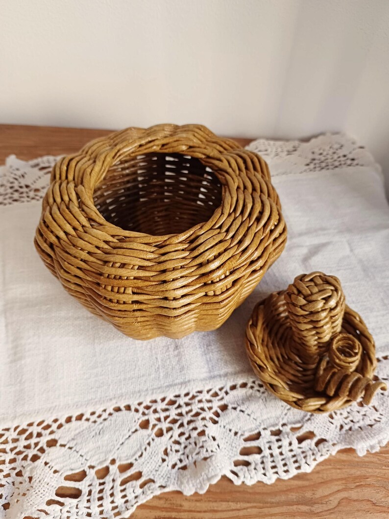 May include: A round, golden-brown woven basket with a pumpkin-like shape and a matching lid. The basket features a textured, coiled weave pattern with a scalloped rim. The separate lid includes a small, central woven handle for easy removal.