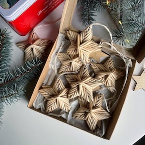 May include: A collection of woven straw star ornaments, some inside a cardboard box with a clear lid. The stars are light brown and have a textured, woven appearance. A red tin with text is visible in the background, along with evergreen branches.