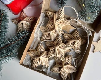 May include: A collection of woven straw star ornaments, some inside a cardboard box with a clear lid. The stars are light brown and have a textured, woven appearance. A red tin with text is visible in the background, along with evergreen branches.