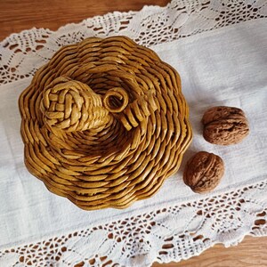 May include: A round, golden-brown woven basket shaped like a pumpkin, featuring a coiled stem on top. The basket is constructed from tightly wound paper or reed material, displaying a textured, circular pattern. Two whole walnuts sit nearby.
