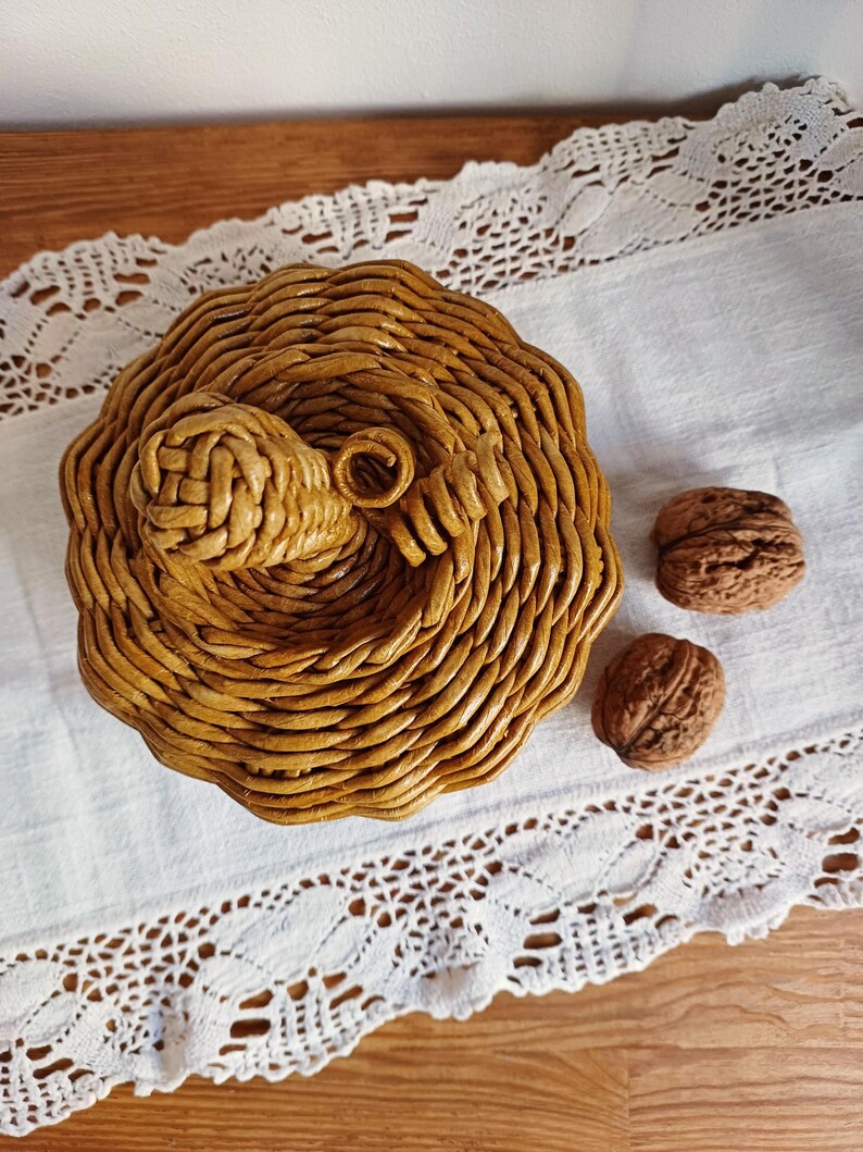 May include: A round, golden-brown woven basket shaped like a pumpkin, featuring a coiled stem on top. The basket is constructed from tightly wound paper or reed material, displaying a textured, circular pattern. Two whole walnuts sit nearby.