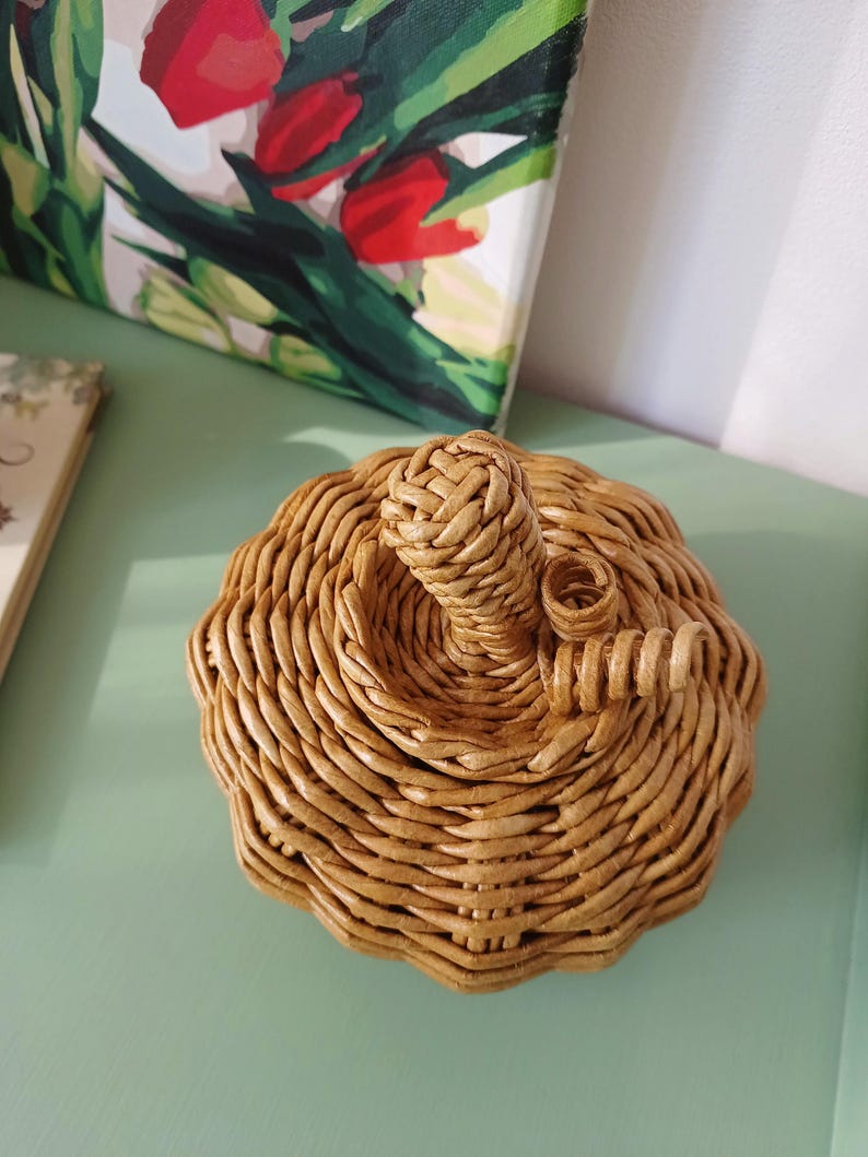 May include: A pumpkin-shaped decorative object crafted from woven light brown material. The item features a rounded, ribbed body with a central stem and a small, coiled vine-like detail extending from the top, showcasing a textured, hand-woven finish.