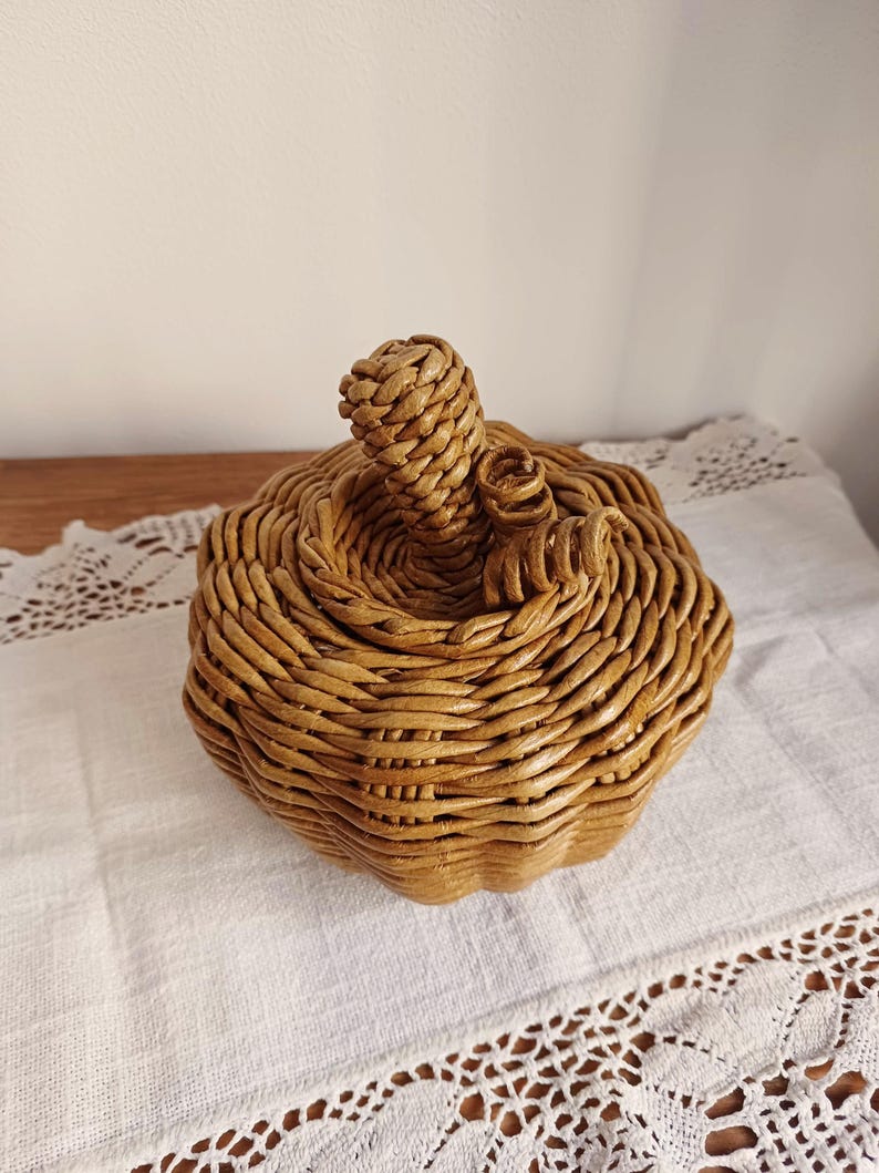 May include: A pumpkin-shaped basket woven from light brown material. The basket features a rounded, ribbed body with a central stem and a small, curled vine detail on top. The woven texture is consistent throughout the entire structure.