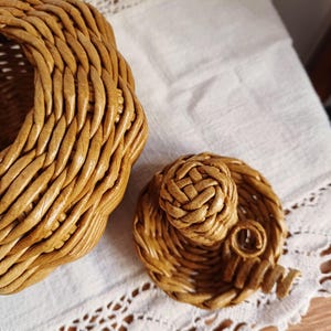 May include: A light brown woven wicker basket with a matching circular lid. The lid features a central woven knob and a small decorative spiral accent. The basket has a thick, braided rim and a textured, hand-woven construction throughout.