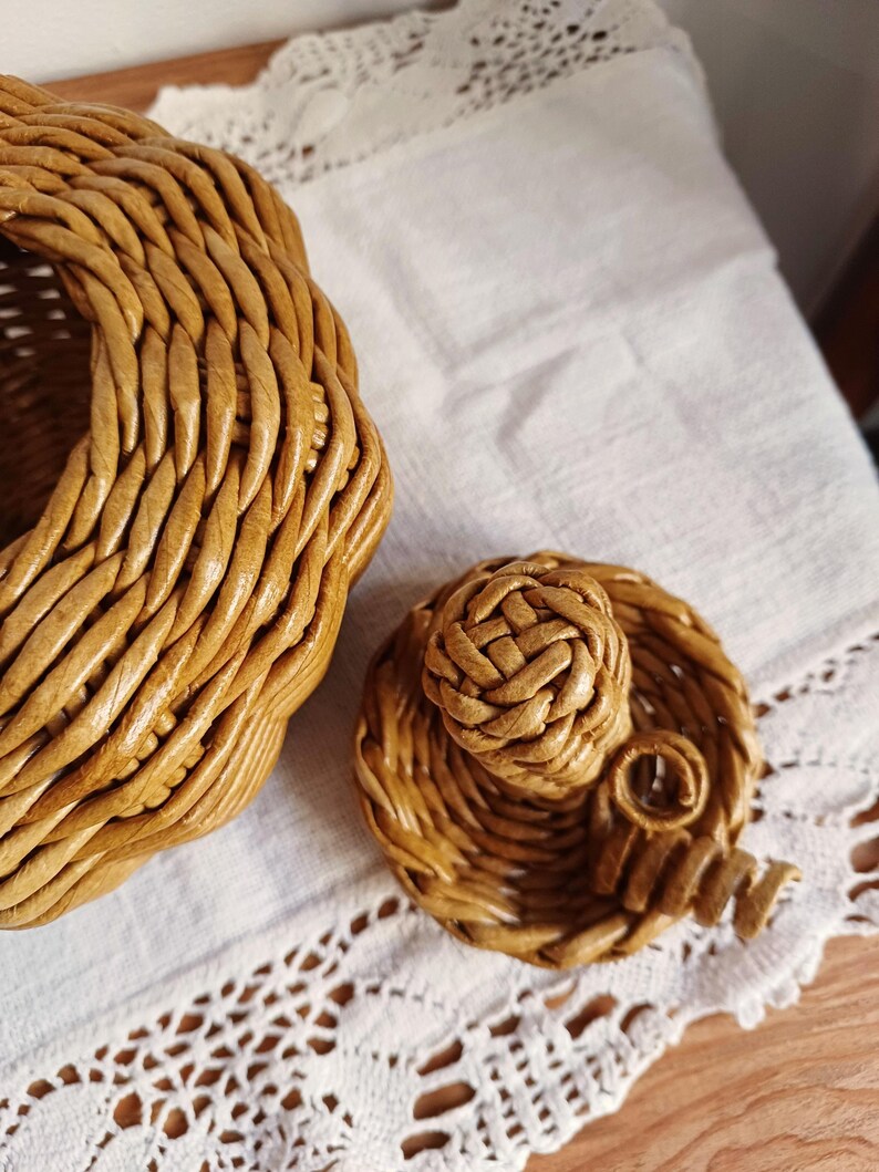 May include: A light brown woven wicker basket with a matching circular lid. The lid features a central woven knob and a small decorative spiral accent. The basket has a thick, braided rim and a textured, hand-woven construction throughout.