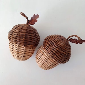 Könnte beinhalten: Zwei eichelförmige Körbe aus geflochtenem Korbgeflecht. Die Körbe sind in Braun- und Beigetönen gehalten, mit einem dunkelbraunen Deckel. Jeder hat ein kleines, braunes, blattförmiges Detail. Die Körbe stehen auf einer weißen Oberfläche.