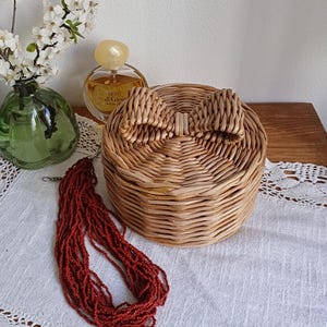 Pode incluir: Uma caixa redonda de vime tecido com uma alça em forma de laço na tampa. Um colar de contas vermelhas e um frasco de perfume estão ao lado da caixa. Um vaso de vidro verde com flores brancas está ao fundo.