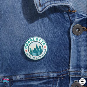 Pu&ograve; includere: Una spilla rotonda con un design turchese e bianco raffigurante lo skyline di Charlotte, Carolina del Nord. La spilla &egrave; attaccata a una giacca di jeans. Il testo "CHARLOTTE NORTH CAROLINA" &egrave; stampato attorno al bordo della spilla.