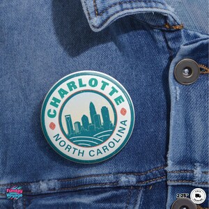 Pu&ograve; includere: Un bottone rotondo con la scritta "CHARLOTTE NORTH CAROLINA" che circonda una grafica dello skyline turchese e bianco. Il bottone &egrave; appuntato su una giacca di jeans blu.