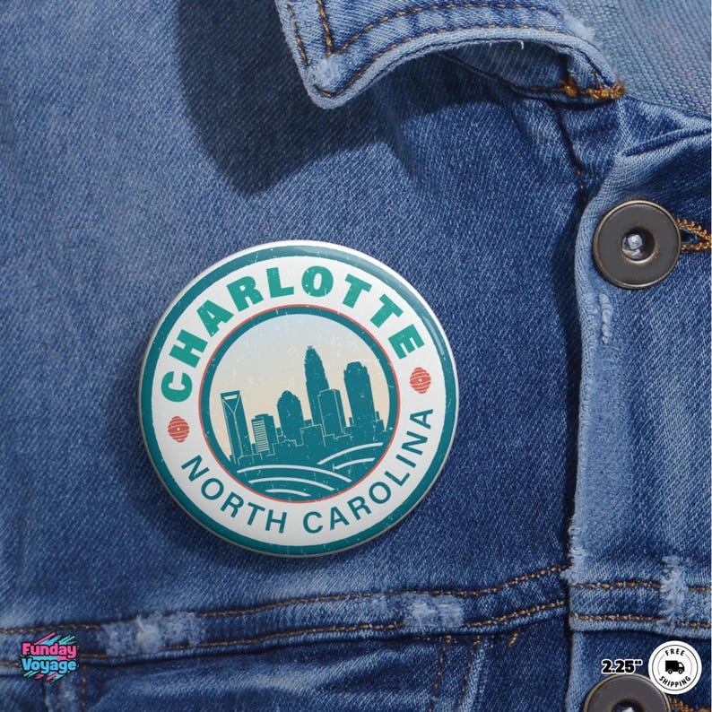 Pu&ograve; includere: Un bottone rotondo con la scritta "CHARLOTTE NORTH CAROLINA" che circonda una grafica dello skyline turchese e bianco. Il bottone &egrave; appuntato su una giacca di jeans blu.