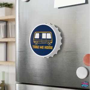 May include: A round bottle cap magnet featuring a dark blue background with a yellow graphic of a camper van and the text "TAKE ME HOME" in yellow capital letters. The magnet has a white scalloped edge and a metallic finish on the sides.