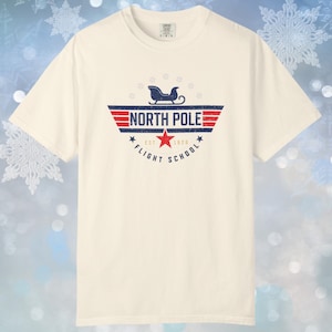 May include: Cream-colored t-shirt featuring a distressed graphic. The graphic includes the text "NORTH POLE FLIGHT SCHOOL EST 1970" with a sleigh and star design. The shirt has short sleeves and a crew neck.