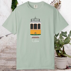 K&ouml;nnte beinhalten: Hellgr&uuml;nes T-Shirt mit einer Illustration einer Stra&szlig;enbahn aus Lissabon. Die Stra&szlig;enbahn ist gelb, orange und wei&szlig;, mit dem Wort "Lissabon" darunter in handschriftlicher Schrift. Das T-Shirt ist aus weichem Material.