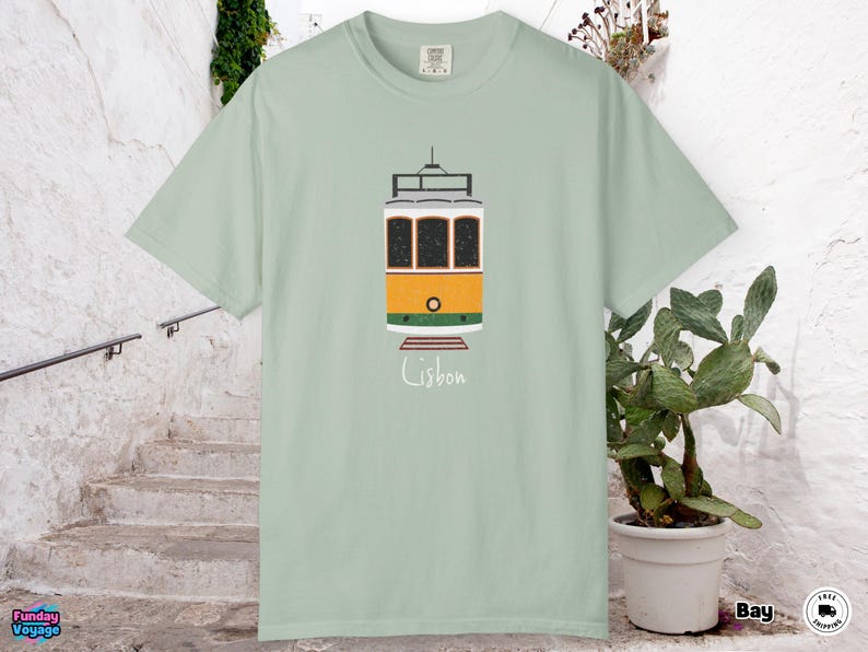 K&ouml;nnte beinhalten: Hellgr&uuml;nes T-Shirt mit einer Illustration einer Stra&szlig;enbahn aus Lissabon. Die Stra&szlig;enbahn ist gelb, orange und wei&szlig;, mit dem Wort "Lissabon" darunter in handschriftlicher Schrift. Das T-Shirt ist aus weichem Material.