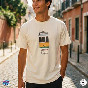 May include: A cream-colored short-sleeve t-shirt featuring a graphic illustration of a yellow and green Lisbon tram. Below the tram, the word Lisbon is printed in a simple black script font. The shirt has a crew neckline and a standard fit.