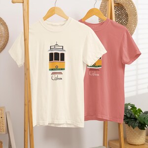K&ouml;nnte beinhalten: Zwei T-Shirts h&auml;ngen an einem Holzst&auml;nder. Eines ist cremefarben mit einer gelb-schwarzen Stra&szlig;enbahngrafik und dem Wort "Lissabon" in Schwarz. Das andere ist einfarbig korallenfarben. Ein Strohhut und Pflanzen sind im Hintergrund.
