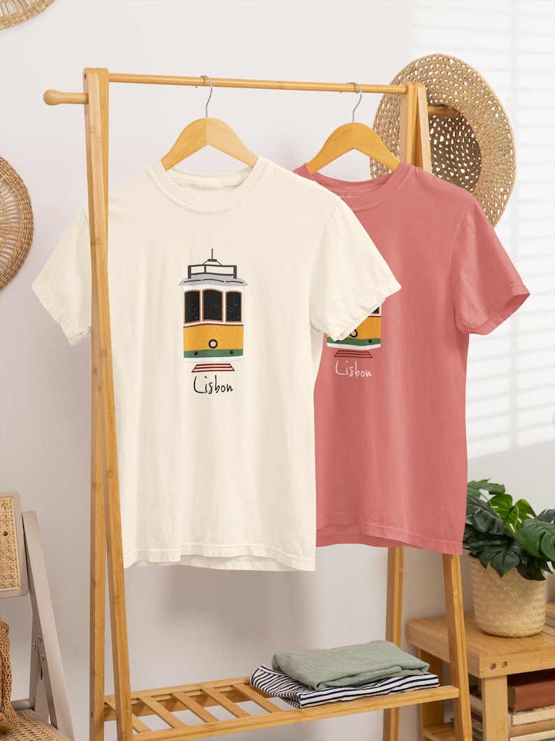 K&ouml;nnte beinhalten: Zwei T-Shirts h&auml;ngen an einem Holzst&auml;nder. Eines ist cremefarben mit einer gelb-schwarzen Stra&szlig;enbahngrafik und dem Wort "Lissabon" in Schwarz. Das andere ist einfarbig korallenfarben. Ein Strohhut und Pflanzen sind im Hintergrund.